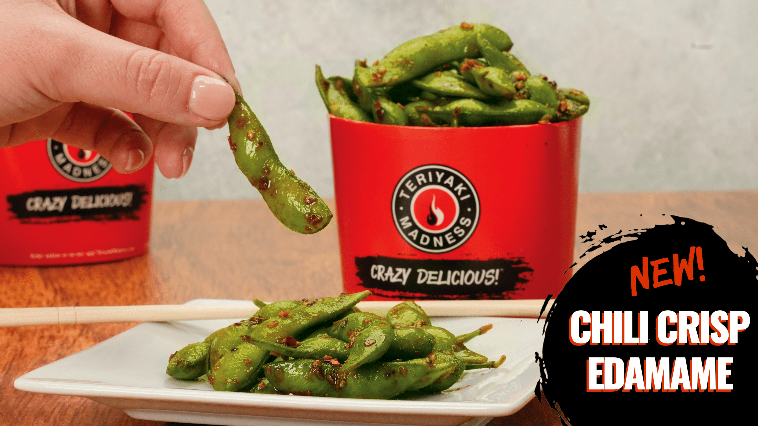 A hand gracefully picking up one of the new, succulent, vibrant, glistening, chili crisp edamame. The light bounces off the sweet little bods with a golden red hue.